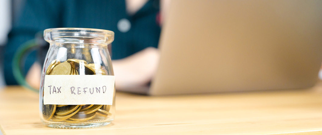 Turn Tax Refund Deposits Into LongTerm Member shutterstock1918967486 | Evok Advertising Jar filled with coins labeled 'Tax Refund' on a wooden desk with a person using a laptop in the blurred background.