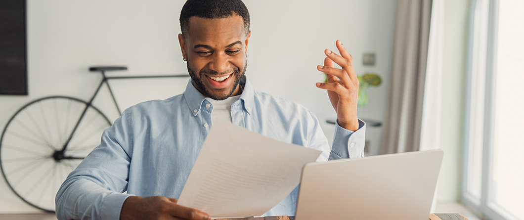 What Are Bigger Tax Refundsshutterstock2739460763 | Evok Advertising Smiling man in a light blue shirt reads a document at a desk with a laptop in a bright home office
