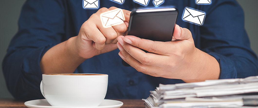 Email Versus SMS | Evok Advertising Person uses a smartphone with floating email icons above a coffee cup on a saucer and a stack of papers sit on a desk nearby