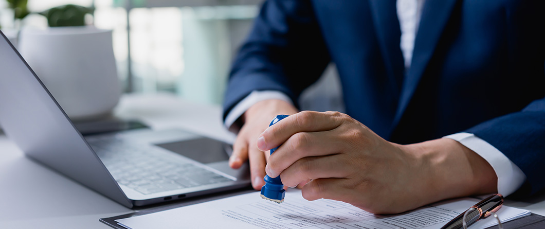 Share Certificate and Money Market | Evok Advertising Person in a suit stamps a document beside an open laptop on a desk signaling approval or signing