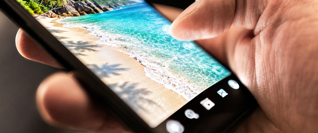 Paid Channel Roles for Destinations | Evok Advertising Close up of a hand holding a smartphone displaying a tropical beach wallpaper with turquoise water and white sand